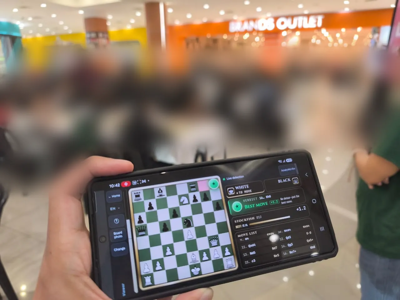 A phone in a shopping-mall tournament showing the live digital board, a “Best move” verdict, Stockfish and the move list; the players in the background are blurred