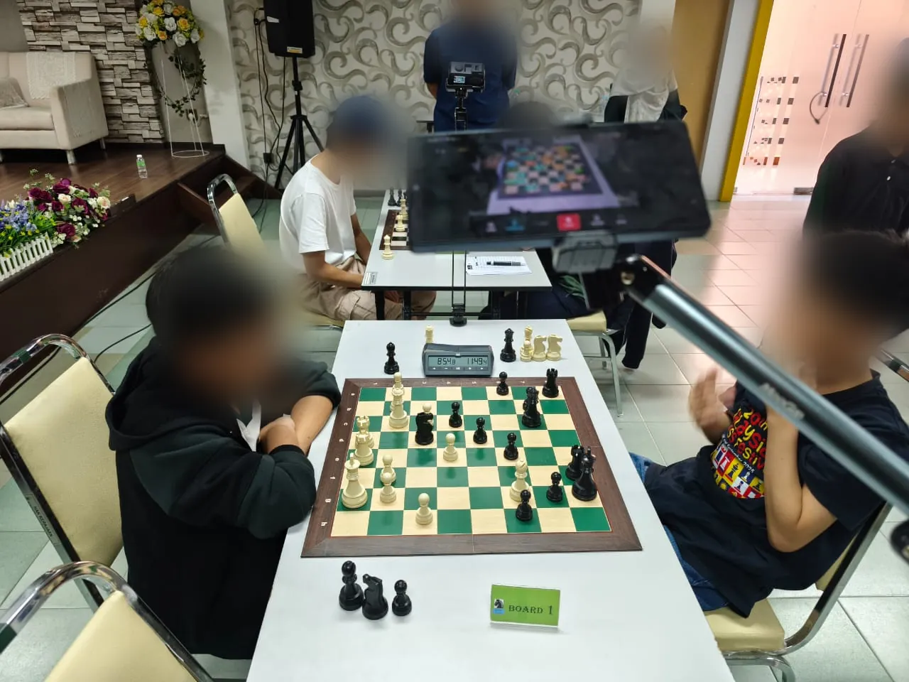 Board 1 of a junior tournament with a phone on an arm filming the board; the players' faces are blurred