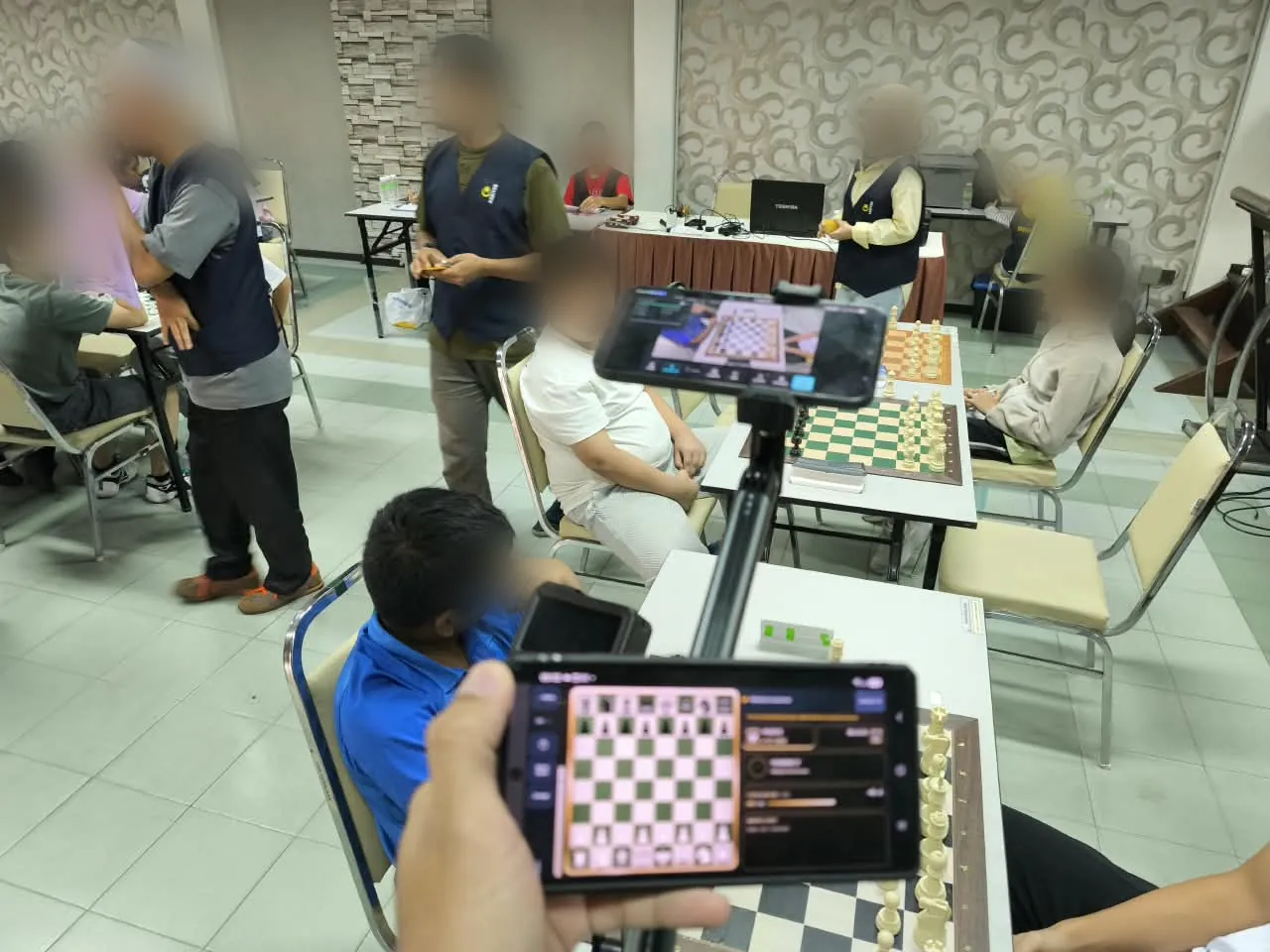 A Viewer phone held in front of a tournament board with the camera phone on a stand behind it; faces are blurred