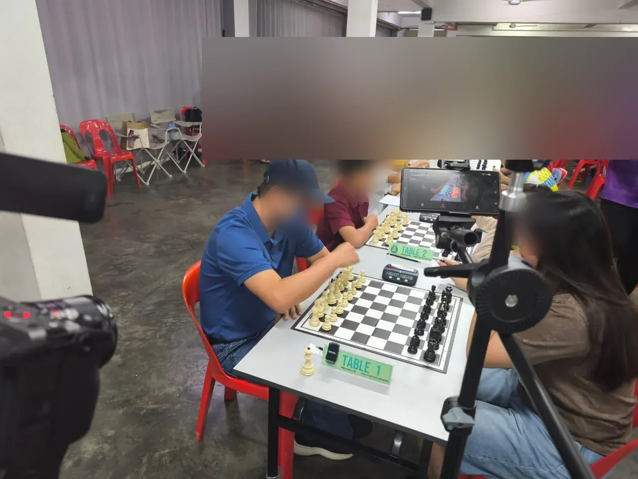 Playing area at the SJKC Keong Hoe tournament with a phone on a stand beside Table 1; faces are blurred