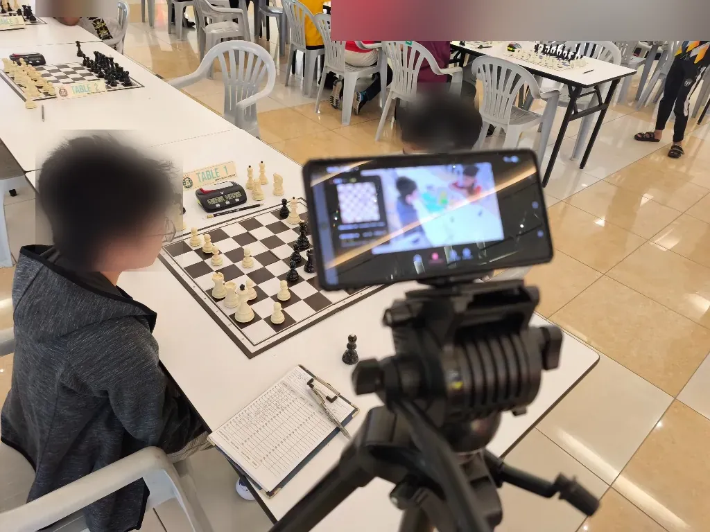 A phone on a tripod at the side of a tournament board, framing the whole board from a stable angle; the players' faces are blurred